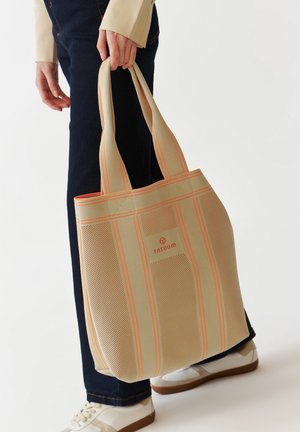 Borsa tote beige con strisce arancioni, realizzata in tessuto a rete. Caratterizzata da doppi manici e una toppa con logo. Indossata da una persona con jeans scuri.