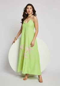 Maxikleid aus hellgrünem Stoff mit pinken, gestickten Palmenblättern, einem V-Ausschnitt und Fransen am Saum. Transparente Schuhe.