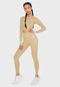 Beige athletischer Crop-Top mit Reißverschluss und passenden hochgeschnittenen Leggings, mit weißen Akzenten und einem Logo an der Hüfte. Weiße Sneaker.