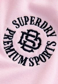 Ružová textúrovaná tkanina s okrúhlym čiernym vyšívaným logom s nápisom "SUPERDRY PREMIUM SPORTS" a prepletenými písmenami "SB."