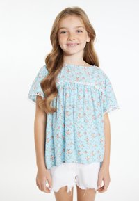 Helles blaues Blumenshirt mit kurzen Ärmeln, Spitzenbesatz und fließendem Design. Kombiniert mit weißen Shorts mit fransigem Saum.