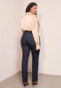 Beige satinblus med långa ärmar, tillsammans med mörkblå höga jeans och bruna klackar. Bakvy som visar sömdetaljer och fickor.