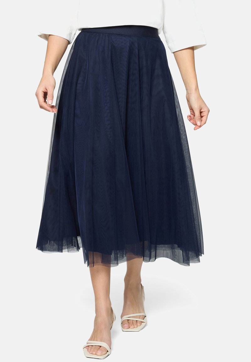 Jupe midi plissée bleu marine avec superposition en tulle, portée avec un haut blanc et des talons beige à bout ouvert sur un fond clair.