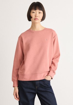 Woman with short dark hair wearing a relaxed pink sweatshirt and dark blue jeans, standing with one hand in pocket against plain background.