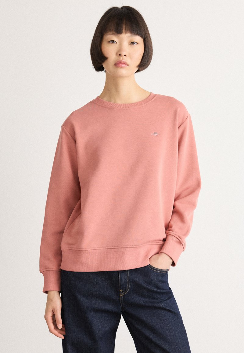 Woman with short dark hair wearing a relaxed pink sweatshirt and dark blue jeans, standing with one hand in pocket against plain background.