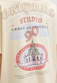 Bežs kokvilnas T-krekls ar daudzkrāsainu grafisko apdruku, kas attēlo ēku, tekstu elementiem dažādās fontu un krāsu kombinācijās, kā arī ovālu fona dizainu.