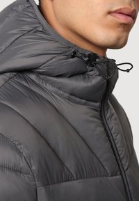 Close-up of a person wearing a dark gray padded jacket with a zip-up front and adjustable drawstring hood.