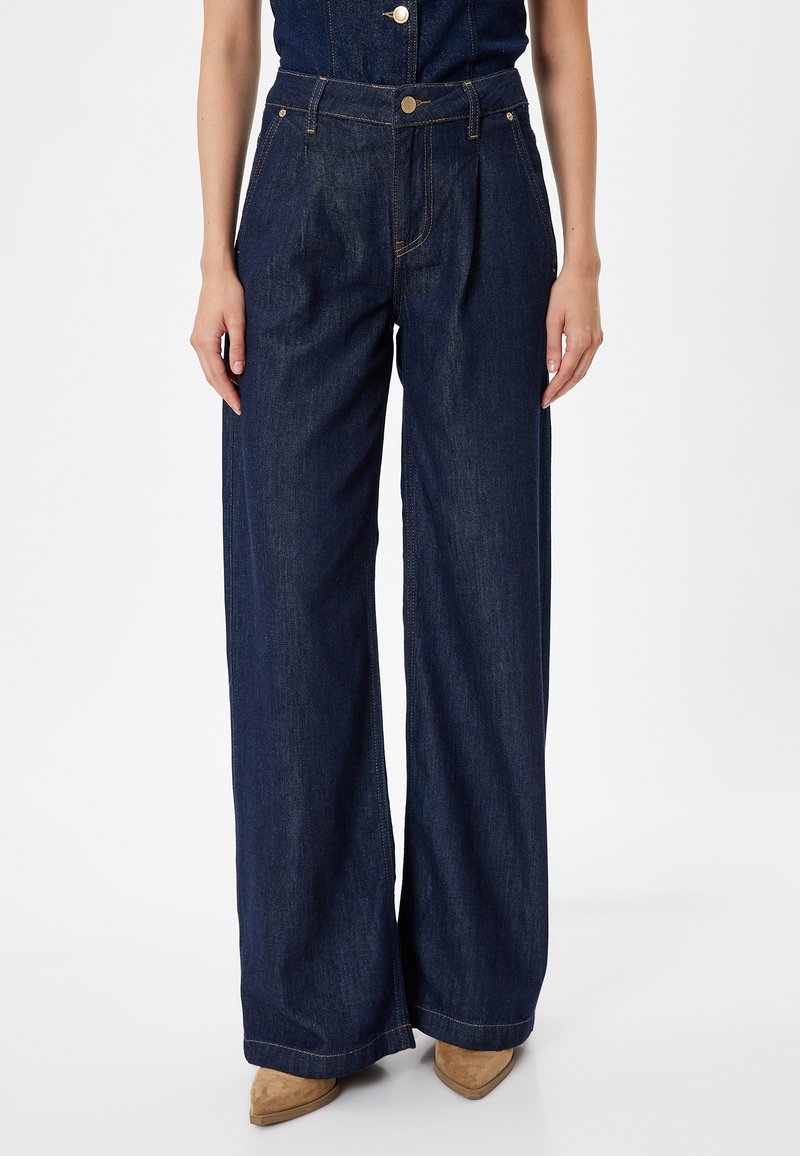 Väljande, mörkblå jeans i denim. Har hög midja, frontveck och sydda detaljer längs sömmarna. Bärs med bruna klackar.