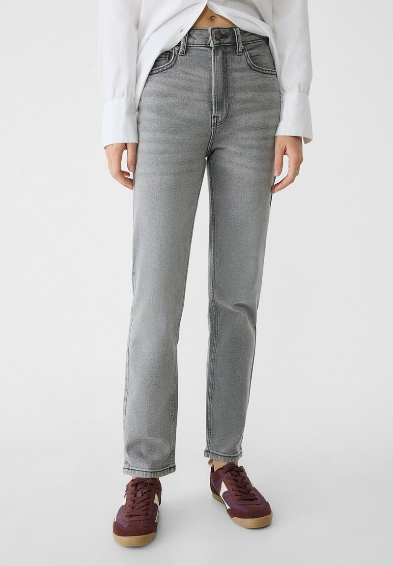 Personne portant un jean gris délavé taille haute, une chemise blanche à larges poignets et des baskets bordeaux avec semelles beiges sur fond blanc.