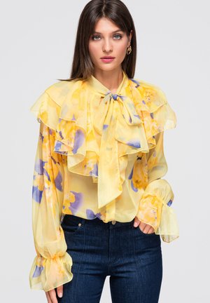 Junge Frau mit glatten braunen Haaren, die eine transparente gelbe Bluse mit Blumenmuster, Rüschen, langen Puffärmeln und einer vorderen Schleife trägt, kombiniert mit dunklen Jeans.