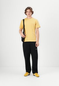 Gul bomulls t-skjorte med korte ermer, kombinert med svarte cargo-bukser med lommer, og gule joggesko. Svart skulderveske.