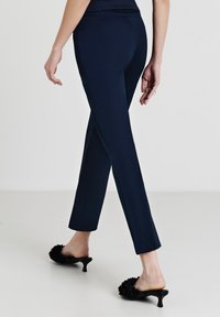 Pantaloni tapered blu navy con una texture liscia, dotati di vita media e tasche posteriori. Indossati con mule nere con tacco che adornano dettagli floreali.