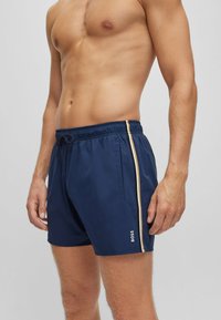 Marineblaue Badehosen mit einem dehnbaren Bund, seitlichen Taschen und kontrastierenden weißen und goldenen Streifen an den Seiten. Mit "BOSS" Branding.