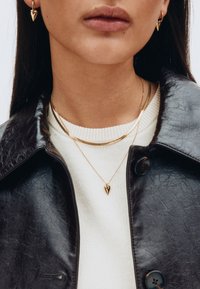 Goldene herzförmige Ohrringe, geschichtete goldene Halsketten, ein weißer gerippter Pullover und eine schwarze Lederjacke mit glänzender Textur.