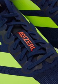 Tengerészkék sportcipő élénk zöld kiegészítőkkel, texturált felső résszel, rugalmas fűzőkkel és piros "ADIZERO" logóval a nyelvén.