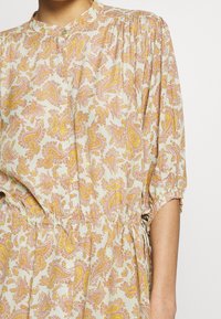 Tiskované šaty z jasně béžové látky s paisley vzorem v růžové a žluté barvě. Nařasený pas a krátké, nabírané rukávy.