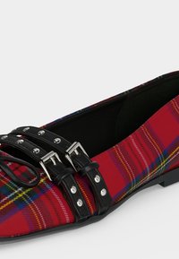 Chaussure plate à carreaux rouges avec doubles brides noires munies de boucles et œillets argentés, bout pointu et semelle basse noire.