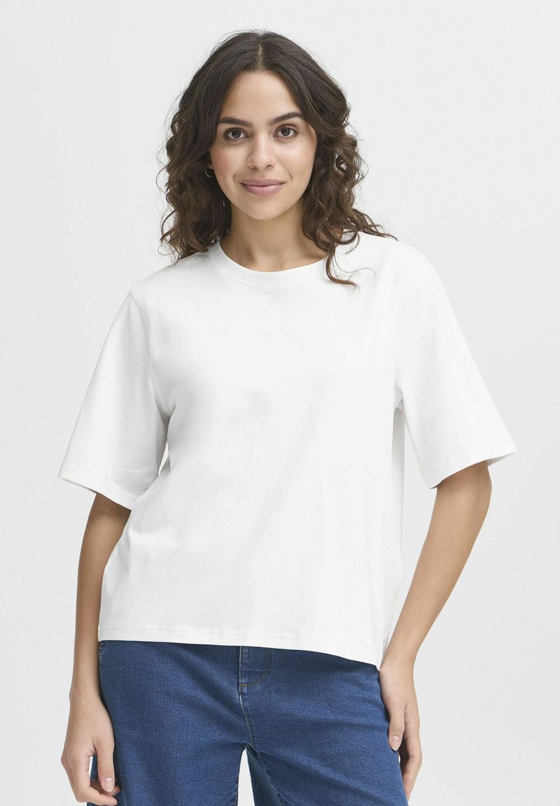 Donna con capelli scuri e mossi, che indossa una semplice t-shirt bianca a maniche corte e jeans blu, in piedi contro uno sfondo grigio chiaro.