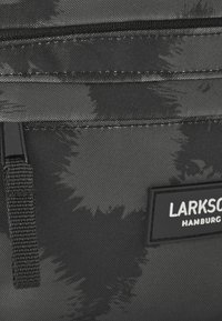 Borsa nera in tessuto con una superficie testurizzata e a motivo. Presenta una cerniera, una tracolla intrecciata e una toppa rettangolare con il marchio e scritte bianche.