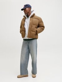 Brun pufferjakke med høy krage over en grå hettegenser, kombinert med lys blå vide jeans og beige sko, satt mot en nøytral bakgrunn.