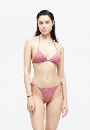 Jeune femme aux cheveux mouillés portant un bikini rose texturé, posant devant un fond blanc uni.