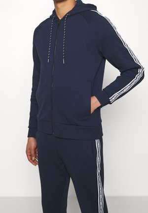 Mężczyzna ubrany w granatową bluzę z suwakiem i pasujące spodnie dresowe z białym paskiem z logo na rękawach i nogawkach, z ręką w kieszeni.