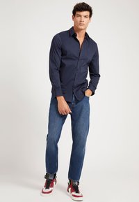 Camisa de botones azul marino de manga larga, combinada con jeans rectos de color azul claro y zapatillas altas rojas y blancas. Diseño sencillo.