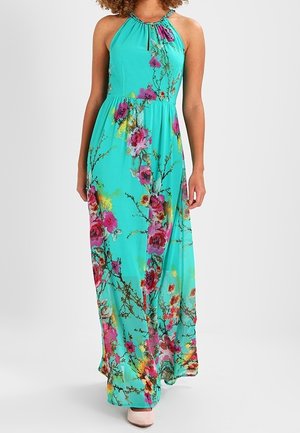 Turkusowa sukienka maxi na szyję z różowo-czerwonym kwiatowym wzorem, noszona przez stojącą osobę na jednolitym tle.