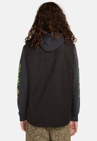 Schwarz vest mit Kapuze, getragen über einem grauen Langarmshirt mit grünen Blumenmustern an den Ärmeln, kombiniert mit olivgrünen Hosen.