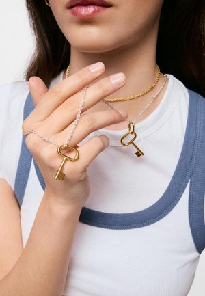 Kobieta nosząca dwa zawieszki w kształcie klucza na srebrnych łańcuszkach i złoty naszyjnik, trzymająca jeden zawieszkę palcami.