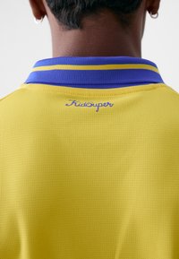 Žuta polo majica s teksturiranim materijalom. Ovratnik ima ljubičaste i žute pruge. "KidSuper" logo izvezen je plavom bojom iznad izreza.