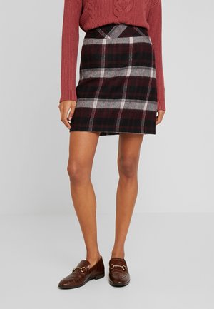 Vrouw die een donkerrode geruite rok, een roestkleurige gebreide trui en bruine loafers met metallic accenten draagt, staand op een eenvoudige grijze vloer.