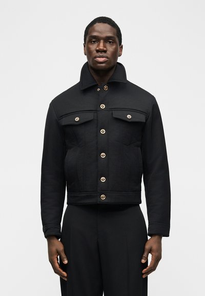 Άνδρας που φοράει μαύρο σακάκι με κουμπιά και δύο τσέπες στο στήθος, στέκεται με τα χέρια χαλαρά απέναντι σε ένα απλό ανοιχτό φόντο.