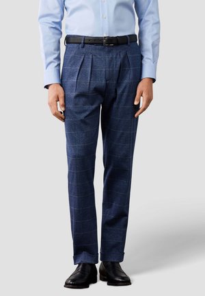 Hombre vestido con pantalones plisados de cuadros azules, cinturón negro, camisa azul claro de manga larga y zapatos de vestir negros, de pie recto.