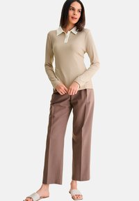 Beige geribde polo met lange mouwen en witte kraag, gecombineerd met bruin wijde broek en lichtgekleurde sandalen. Soepele texturen, casual ontwerp.