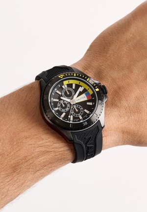 Crni Nautica ručni sat sa žutim detaljima, tri podbrojčanika i crnim teksturiranim gumenim remenom na zapešću osobe.