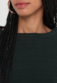 Top de textura verde oscuro con rayas horizontales, que presenta un escote amplio y acentos de cabello trenzado. Pendientes grandes de aro dorado.