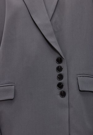 Blazer gris à texture lisse, avec un revers cranté, six boutons noirs et deux poches latérales. Coupe ajustée avec une forme structurée.