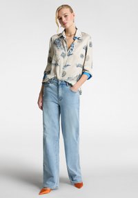 Camicetta beige chiaro con motivo grafico blu, maniche lunghe arrotolate, abbinata a jeans a gamba larga blu chiaro e tacchi a punta arancioni.
