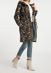 Parka de estampado camuflaje en verde oscuro y gris, con capucha ajustable, cintura con cordón, dos bolsillos frontales y cierre de cremallera. Lleva puesta sobre unos vaqueros azul y botas de tobillo beige.