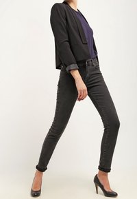 Blazer noir court porté sur un haut bleu marine, associé à un jean skinny noir et des talons hauts noirs. Tissu lisse avec revers de manches roulés et coupe ajustée.