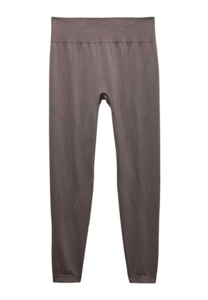 Kobieta w dopasowanym sportowym staniku w kolorze taupe i legginsach z wysokim stanem, białe trampki, z bluzą narzuconą na ramię, klęcząca na jedno kolano.