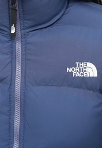 Kék párnázott dzseki eleje függőleges cipzárral, a jobb mellkasi részén fehér hímzett "The North Face" logóval.