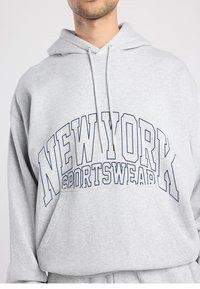 Grijze hoodie met een voorzak, voorzien van donkerblauwe tekst "NEW YORK SPORTSWEAR" in vetgedrukte letters over de borst. Zacht materiaal.