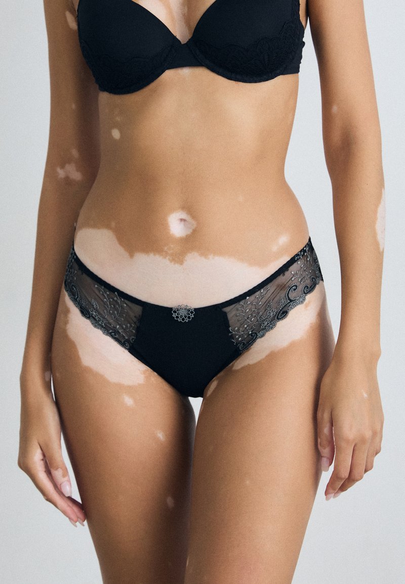Zwarte kanten en stoffen lingerie met ingewikkelde borduurdetails, een middenhoogte snit en een decoratief centraal detail met gekartelde randen.