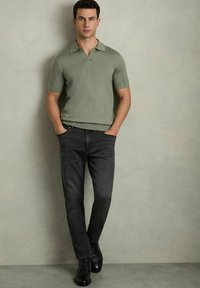 Polo din bumbac de culoare verde deschis, cu mânecă scurtă și guler plat, purtat cu blugi gri închis și pantofi negri cu șireturi, stând în fața unui fundal neutru.