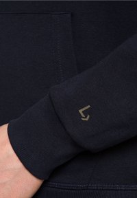 Námořnická mikina s jemnou texturou, se zlatým logem na manžetě a přední kapsou. Detailní záběr na rukáv a logo.