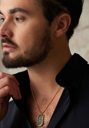 Gold dog tag pendant with engraved design on a gold chain, paired with a gold hoop earring. Black shirt with an open collar.