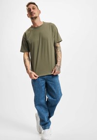 Camiseta de algodón verde oliva de manga corta, con cuello redondo y un ajuste relajado, combinada con jeans holgados azules y zapatillas blancas.