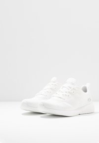 Ett par vita Skechers löparskor med texturerad stickad tyg och gummisålar på en vit yta och bakgrund.